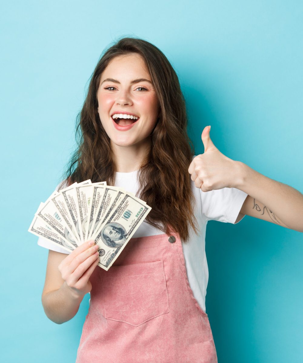 Smiling happy woman holding money, dollar bills and showing thumb up, recommending fast cash loan and looking satisfied, standing over blue background.